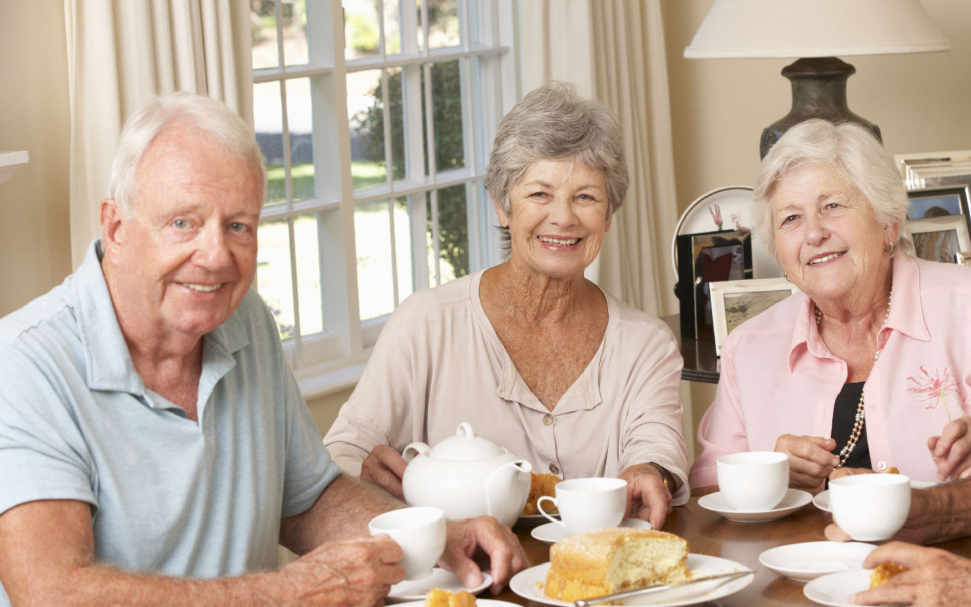 Two broadly smiling senior women and a man having a cake and coffee.