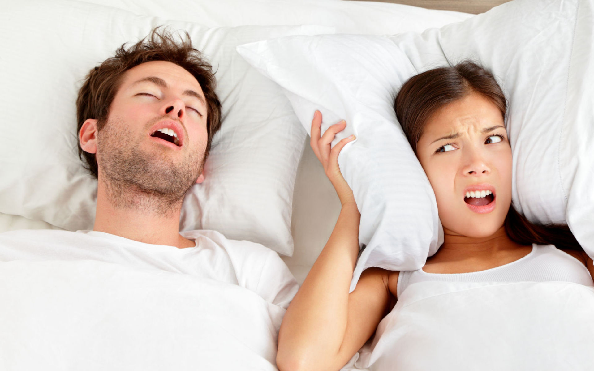 An annoyed woman covering ears with a pillow due to her partner's snoring.
