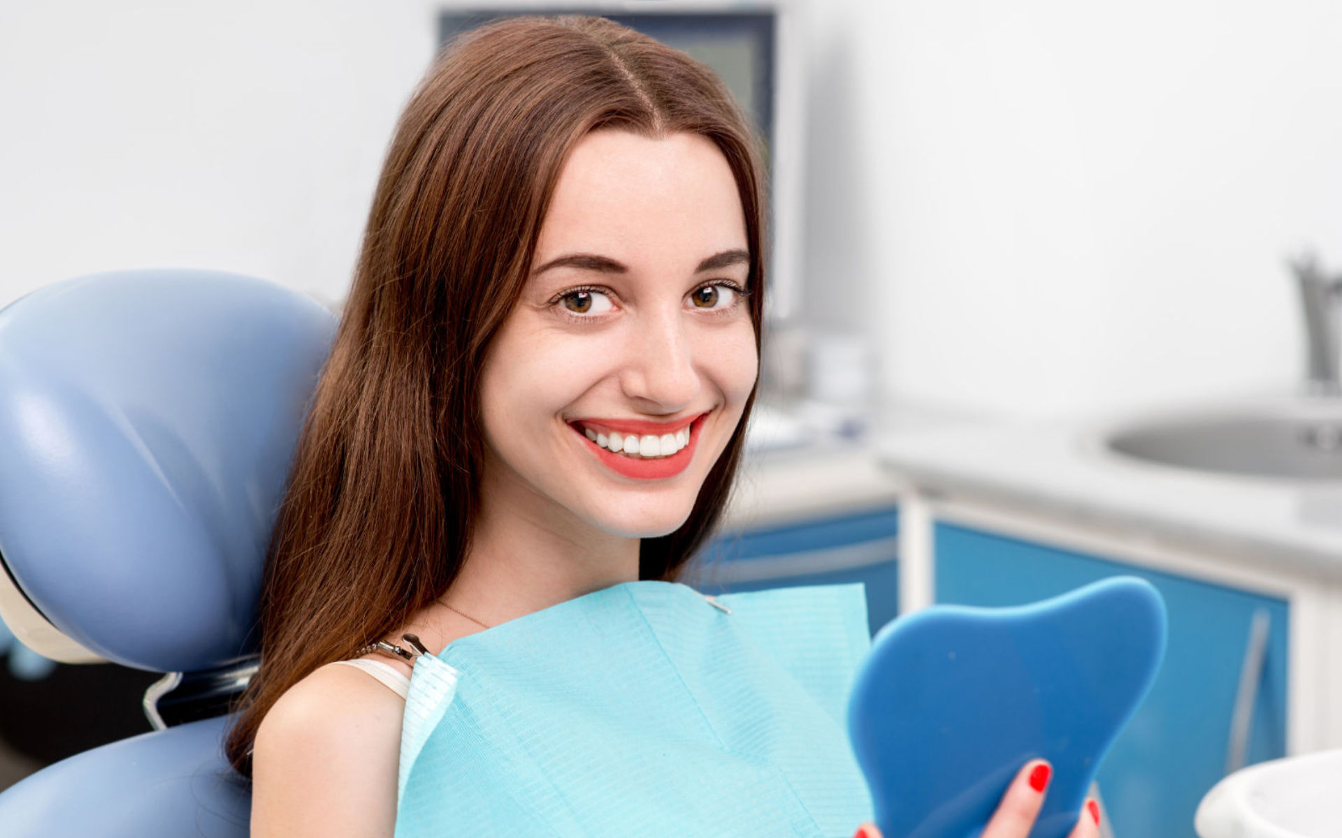 A satisfied young woman sitting in a dentist chair and looking at her beautiful teeth in a mirror.