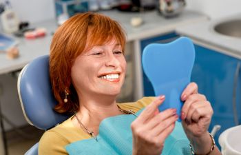 Smiling mature woman in a dental chair looking at her teeth after restorative treatment in a mirror.